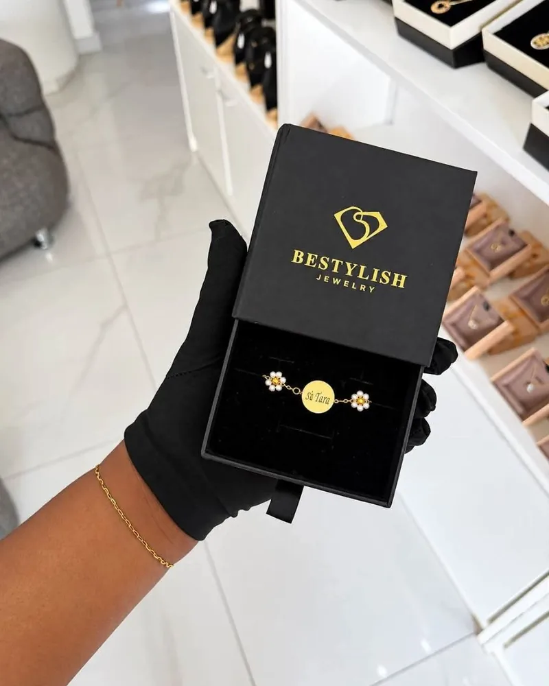 Main gantée présentant un écrin Bestylish avec bracelet fleurs et médaille